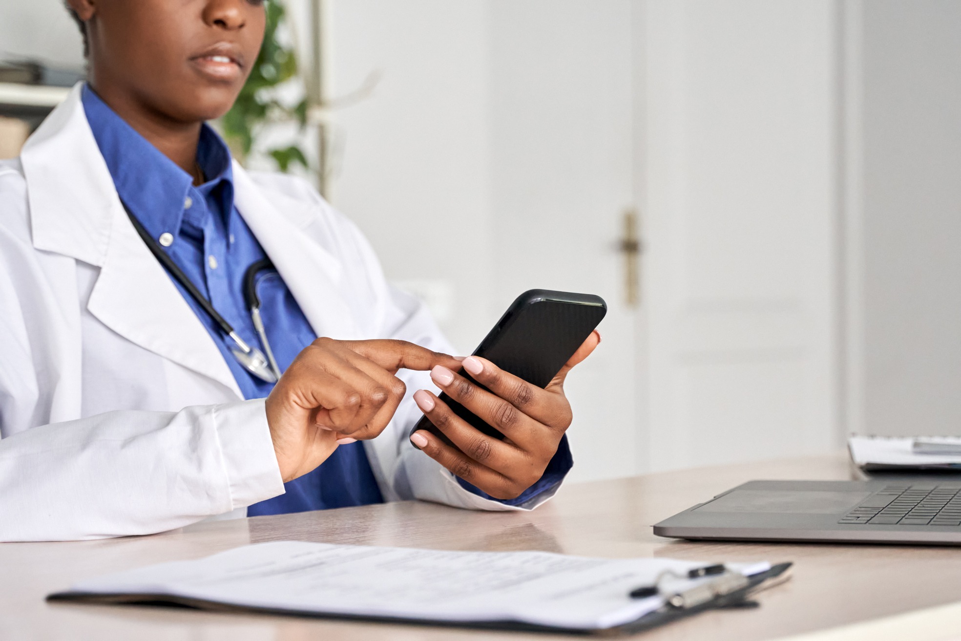 A healthcare professional in a white coat uses a smartphone while seated at a desk with a laptop and paperwork, illustrating modern patient care.