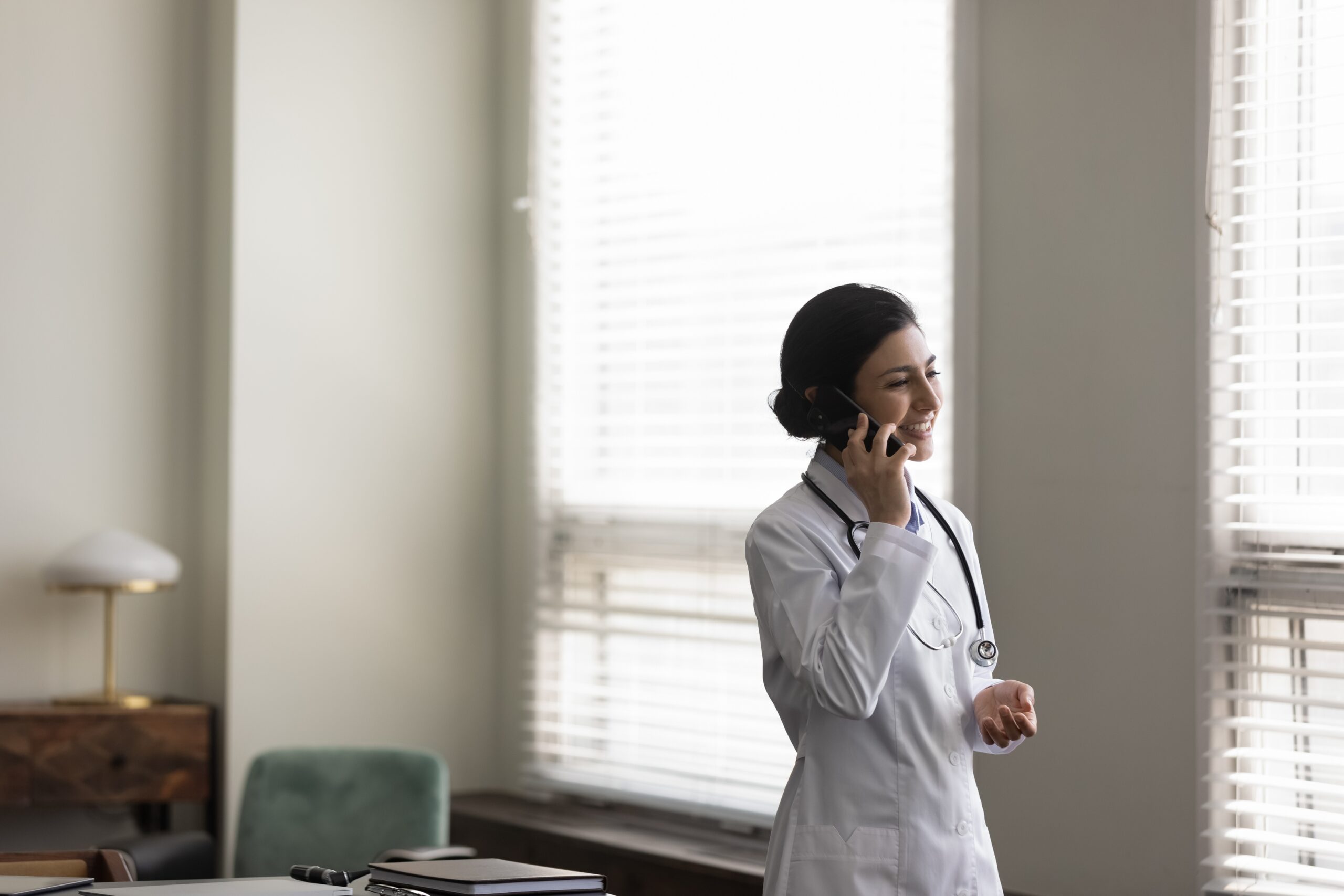 A smiling doctor wearing a white coat and stethoscope speaks on the phone in a well-lit office, highlighting the importance of telehealth communication.