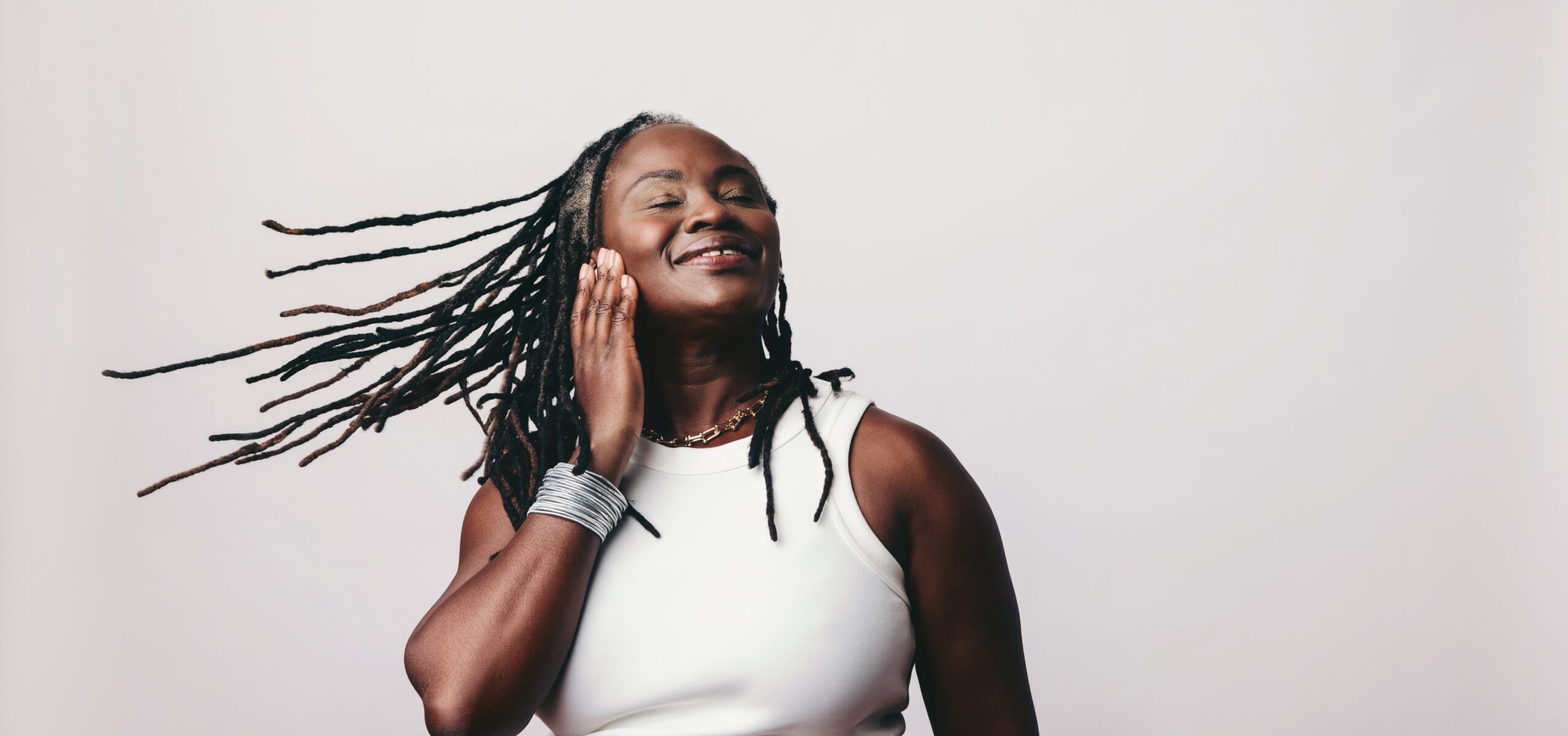 A joyful woman with long dreadlocks, wearing a sleeveless white top, smiles while gently touching her face against flowing hair, set against a soft pink background.